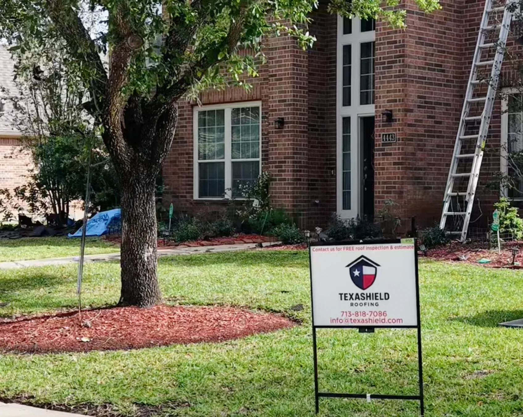 TexaShield Roofing yard sign outside a brick home with a ladder at the roofline.